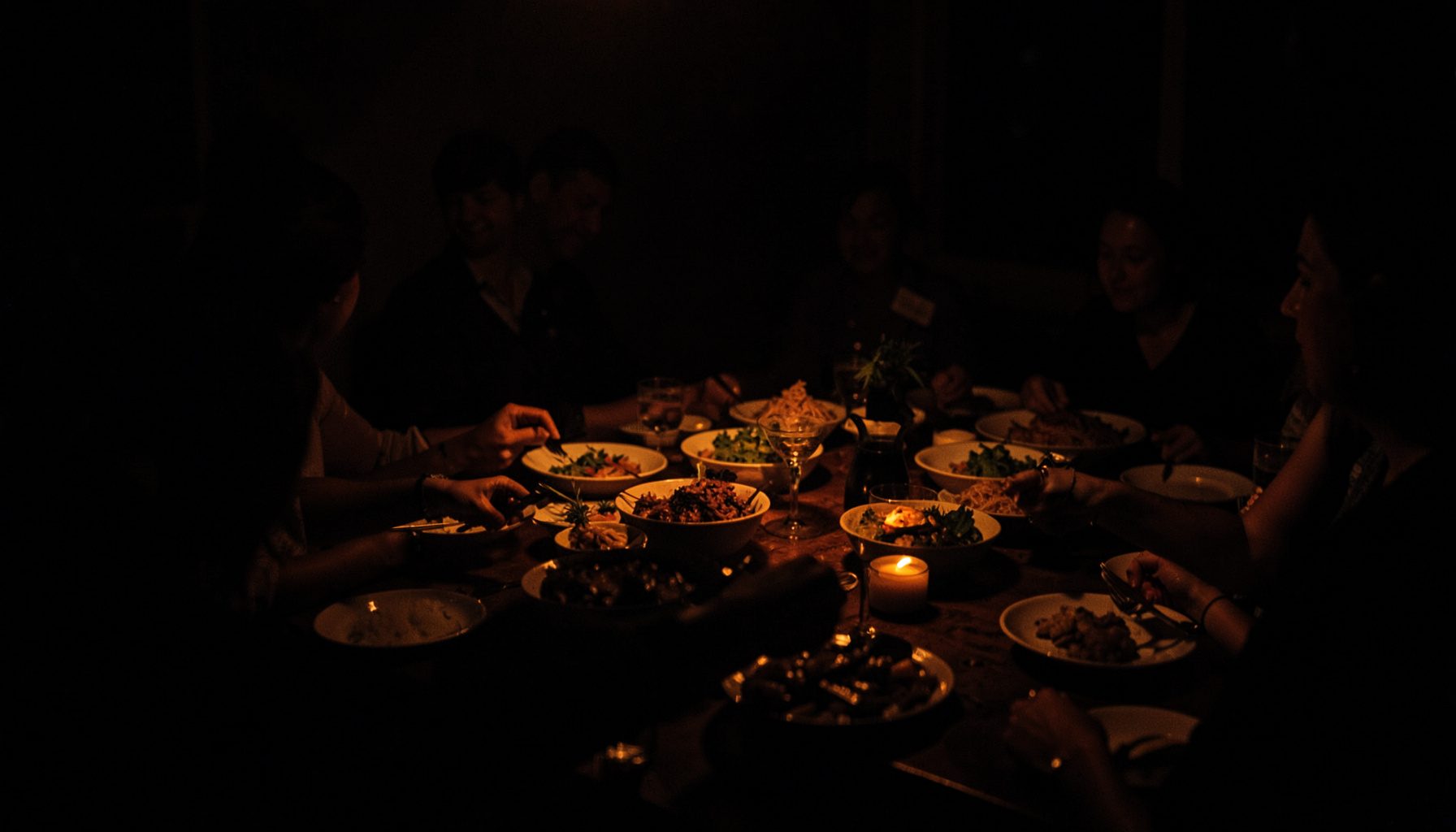 Dark, low-key photograph of a shared community meal table, deep shadow with warm amber highlights