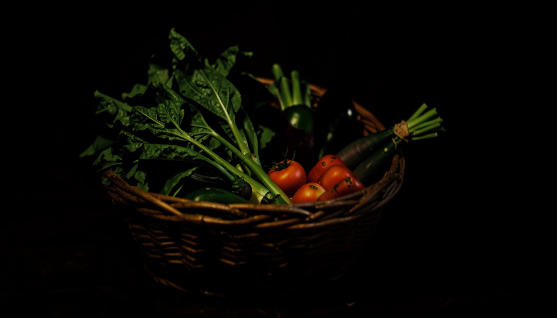 Dark moody photograph of fresh vegetables in a woven basket, deep greens and earthy browns