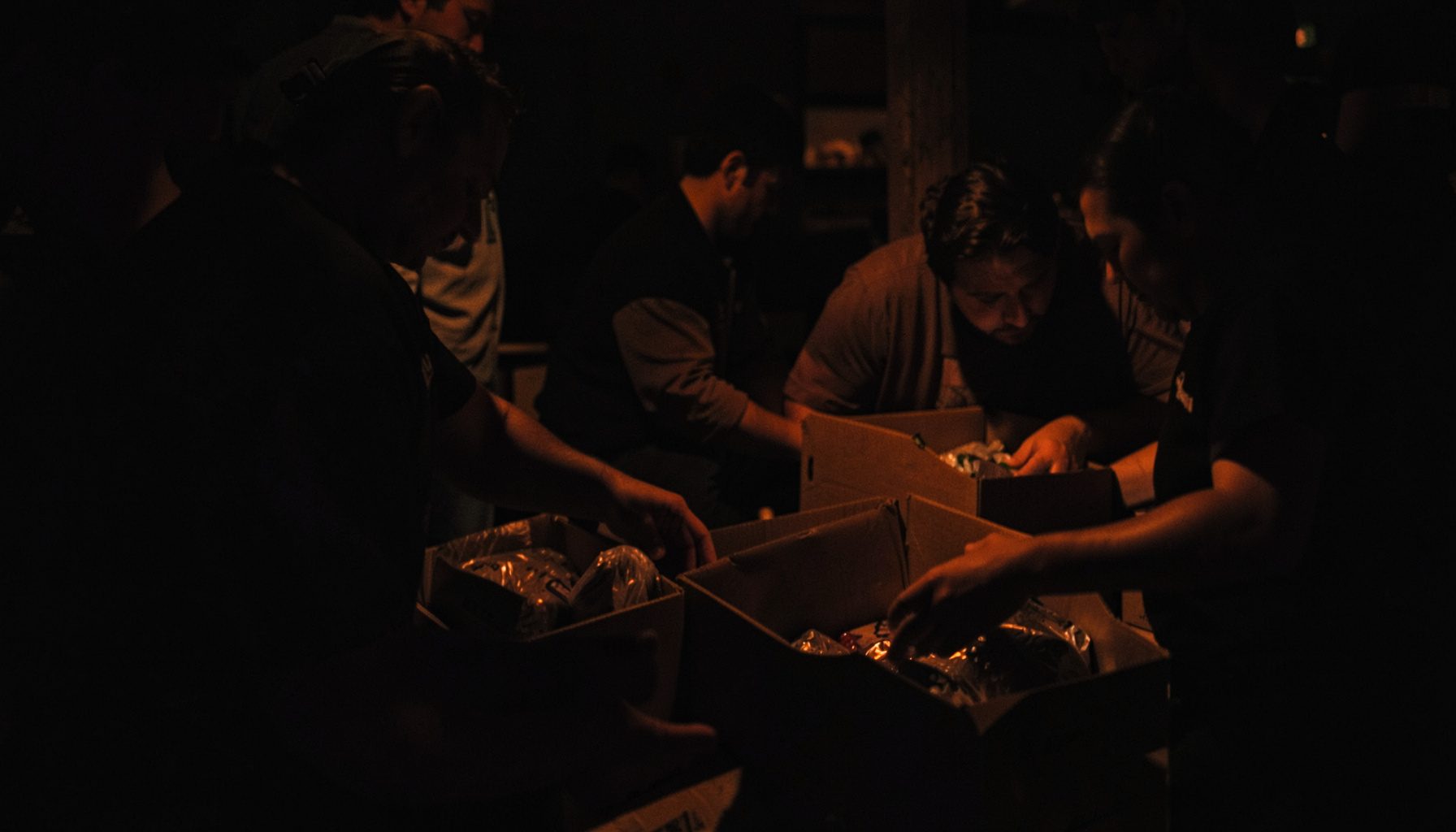 Moody low-light photograph of volunteers packing food boxes, warm skin tones against dark background